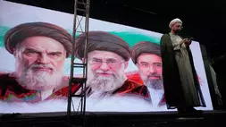 Seorang ulama memeriksa ponselnya di atas panggung di depan layar yang menampilkan potret mendiang pendiri revolusi Iran Ayatollah Khomeini (kiri), mendiang Pemimpin Tertinggi Ayatollah Ali Khamenei (tengah), dan Pemimpin Tertinggi Ayatollah Mojtaba Khamenei, selama pertemuan pro-pemerintah di Teheran, Iran, Minggu (7/6/2026). (Foto: AP/ Vahid Salemi)