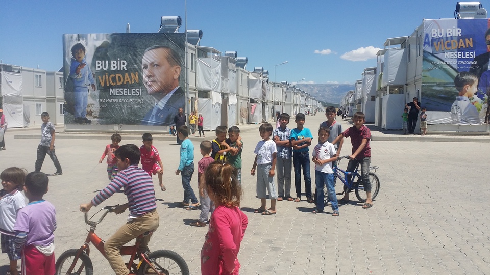 Refugee children at the Kahramanmaraş camp stare and play in front of the large banner at the back featuring a distressed child facing Turkish President Recep Tayyip Erdoğan and reads: “It is a matter of conscience”. (Javier Delgado Rivera)