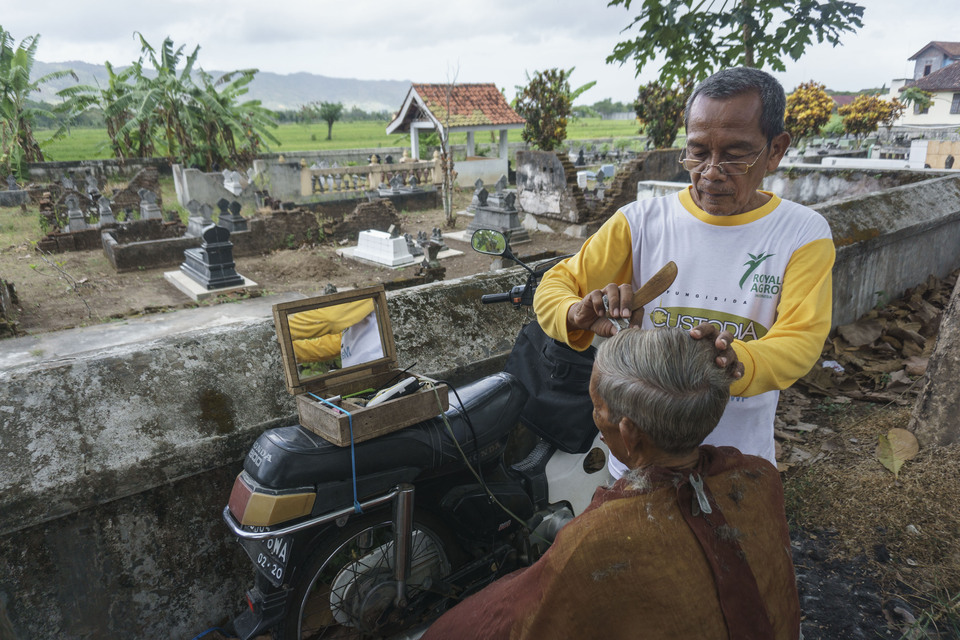 Suradi, 67, cuts the hair of his customer in front of the Klisat village cemetery in Srihardono, Pundong, Bantul, Yogyakarta, on Wednesday (02/08). The barber works five days a week and leaves it up to his clients how much they want to pay. (Antara Photo/Hendra Nurdiyansyah)