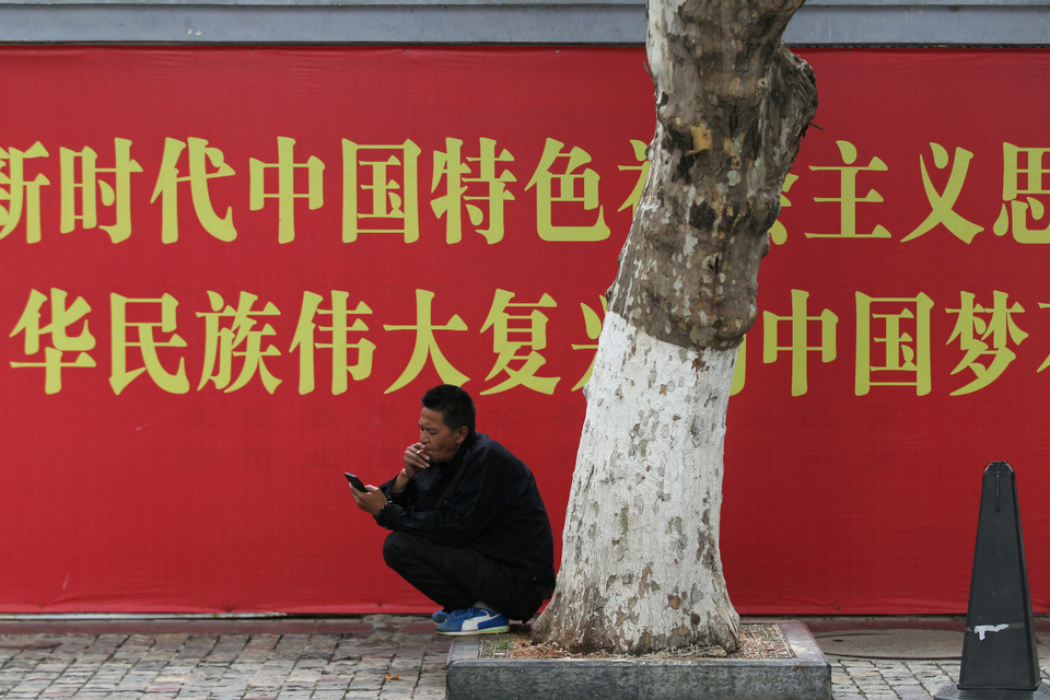 A man smokes a cigarette while looking at his mobile phone in front of a propaganda banner in Kunming, Yunnan province, China, Wednesday (30/05). (Reuters Photo/Wong Campion)