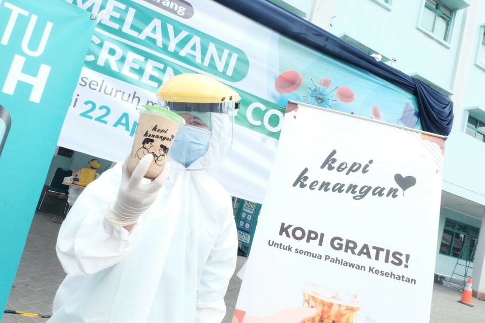 This 2020 file photo shows a person wearing personal protective gear holding a cup of Kopi Kenangan's coffee and standing beside a placard saying free coffee for Indonesian healthcare workers. (Photo courtesy of Kopi Kenangan)