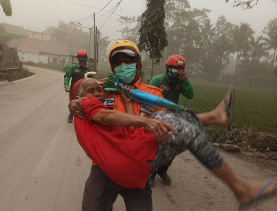 A rescue worker carries an old woman on the slope of Mt. Semeru in the East Java District of Lumajang as the volcano erupts and sends hot ashes into the air on December 4, 2022. (Photo courtesy of the National Disaster Mitigation Agency/BNPB)