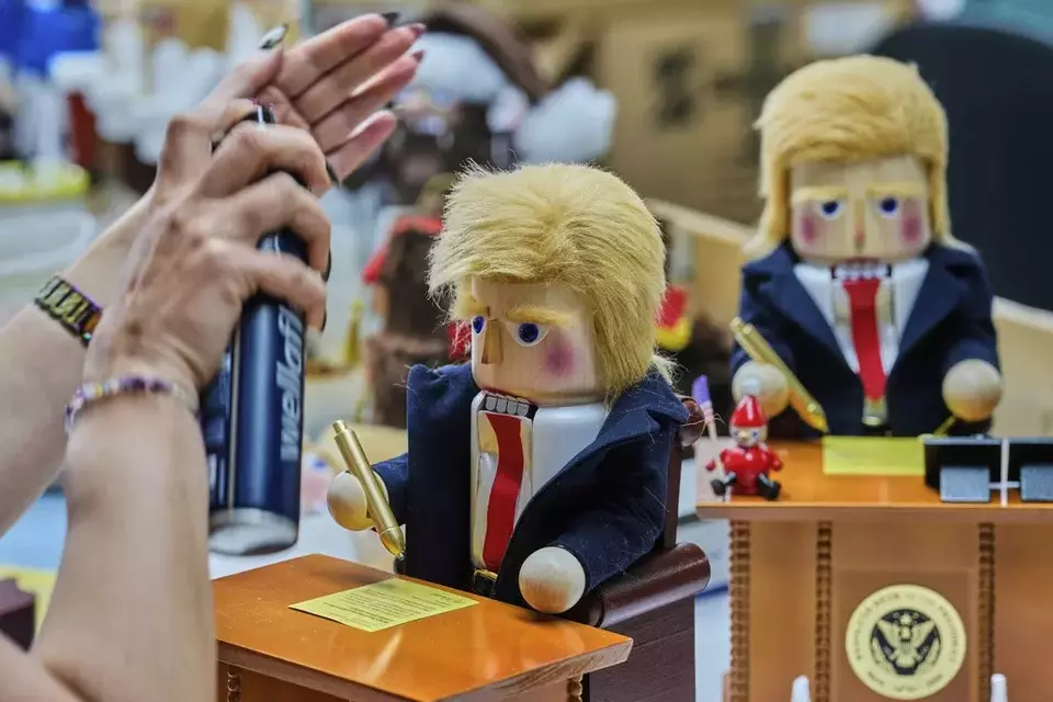 A woman uses hairspray to fixe the hair on a nutcracker titled 'Resolute Desk of the President,' featuring a seated figure signing a 'presidential proclamation,' at the Steinbach-Volkskunst nutcracker factory in Marienberg, Germany, Tuesday, April 15, 2025. (AP Photo/Markus Schreiber)