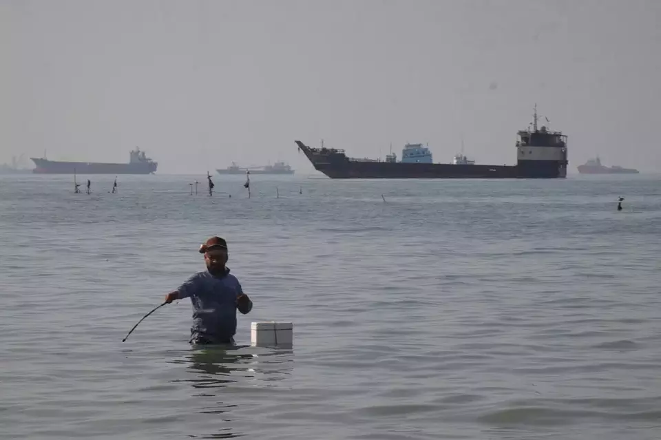 A man stands in the water, appearing to fish, as bulk carriers, cargo ships, and service vessels line the horizon in the Strait of Hormuz off Bandar Abbas, Iran, Monday, April 27, 2026.(Razieh Poudat/ISNA via AP)

